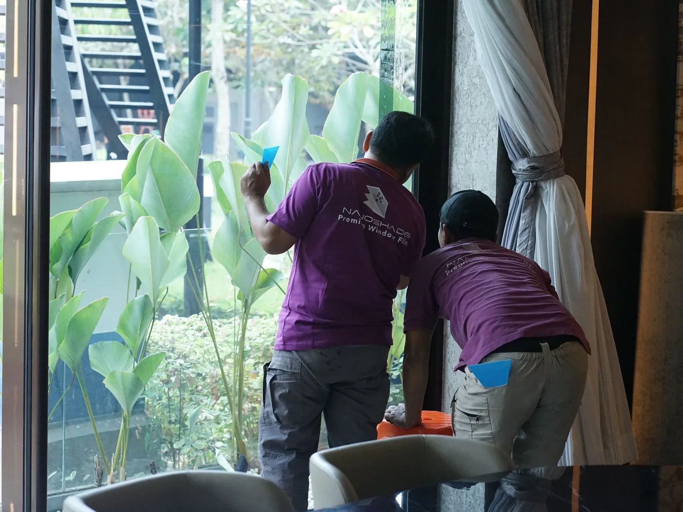 Dua teknisi NanoShade berseragam mengerjakan jendela tinggi di sebuah rumah, tanaman taman terlihat di luar dan gorden di satu sisi