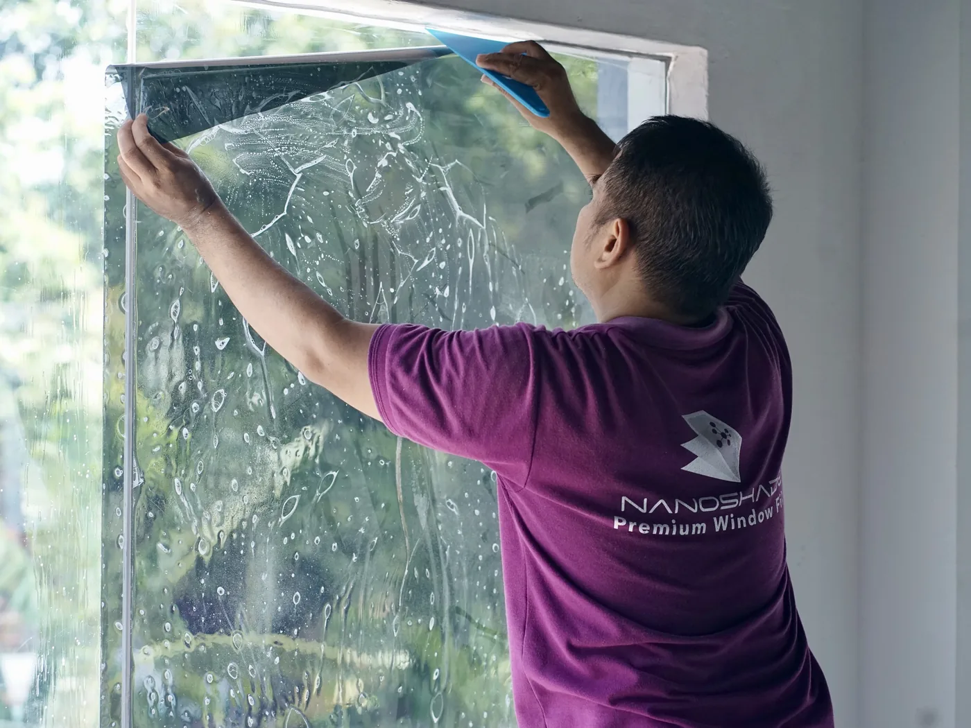 Seorang teknisi NanoShade meratakan lembaran film pada kaca basah dengan squeegee biru, tanaman hijau terlihat melalui kaca
