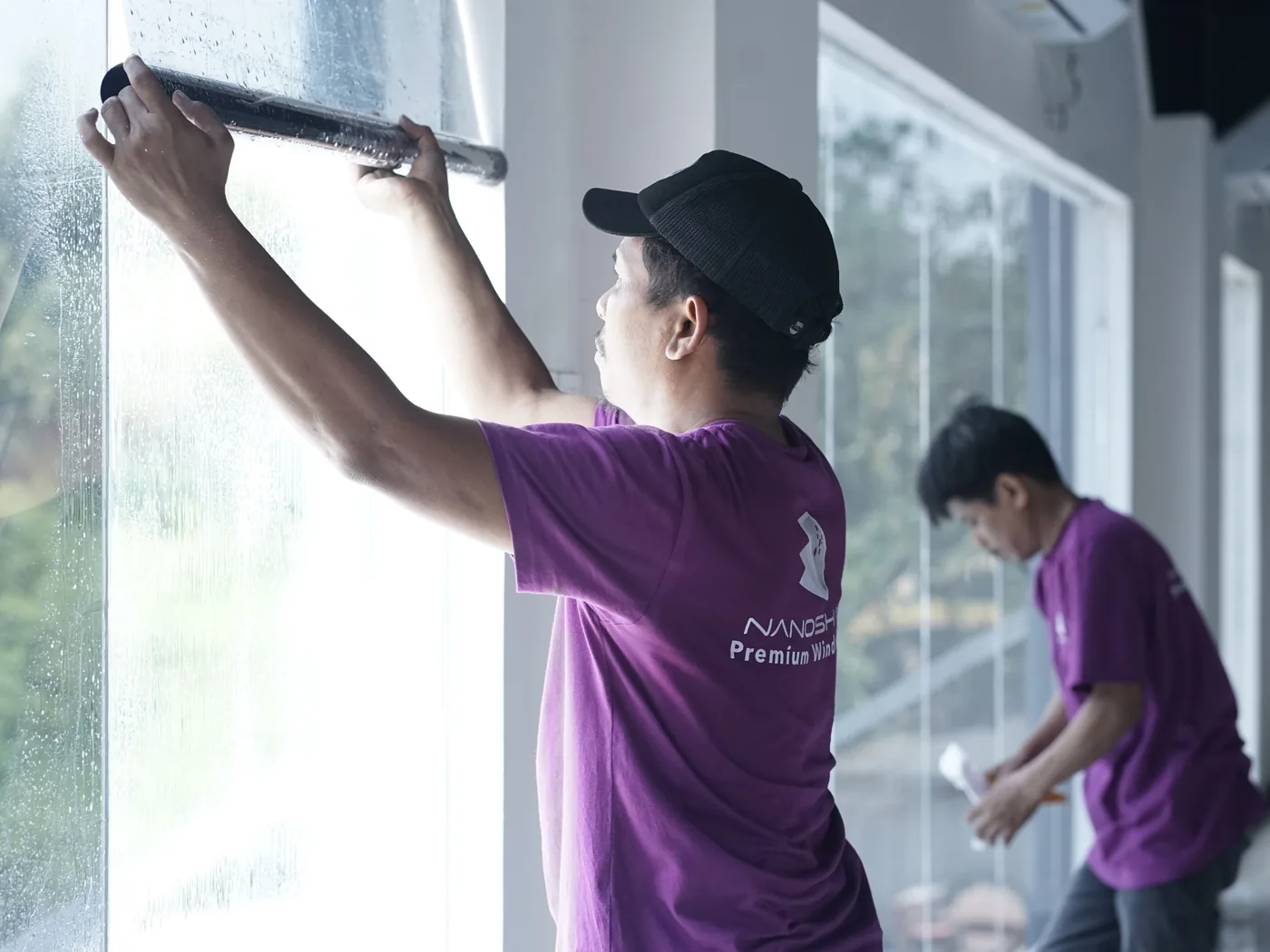 Dua teknisi NanoShade berseragam menarik squeegee di atas jendela tinggi yang basah di gedung lapangan padel